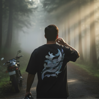Man standing in a forest with a motorcycle, wearing a black t-shirt with a white design.